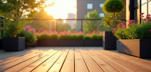 Obraz premium Rooftop balcony with green plants and pink flowers at sunset. Wooden deck reflects warm sunlight. Modern building facade visible in background city skyline.