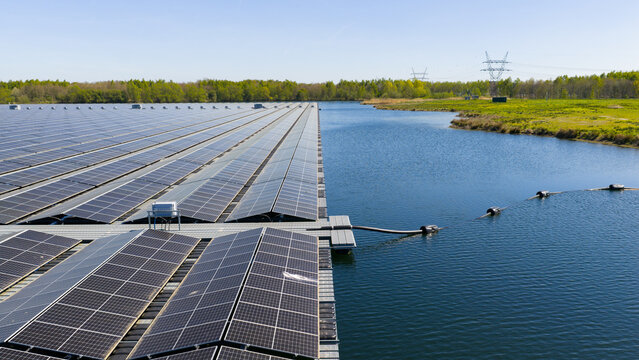 Aerial view of a large floating solar farm on a calm lake surrounded by green fields and lush trees under a clear blue sky Zwolle, Overijssel, Netherlands.
