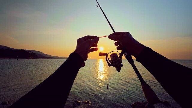 First person view of hands preparing fishing tackle and lure on spinning rod against sunset over sea, detailed setup before casting