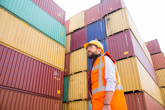 Man in yellow hard hat and orange safety vest stands beside stacked shipping containers in a container yard