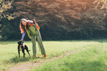 Naklejka premium Happy woman wearing backpack and using trekking poles, petting her dog on a green grass outdoor trail, enjoying companionship