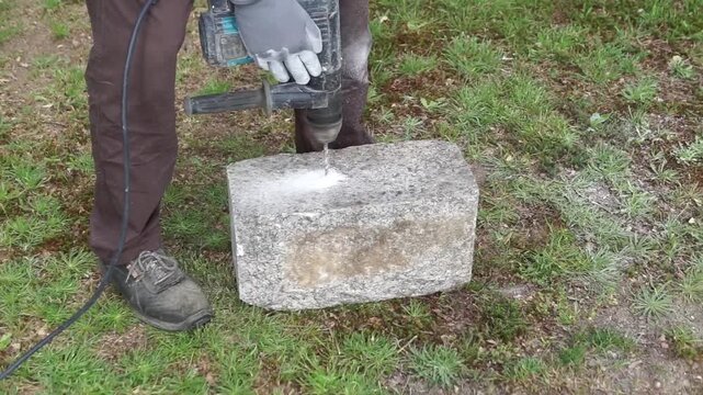 A professional using a hammer drill to create a hole in a large stone block, with dust flying during the process.