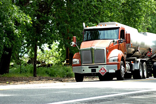 Oncoming fuel tanker on two-lane rural highway with trees in background.