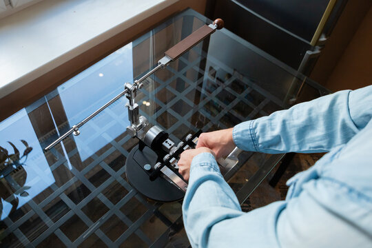 Man adjusting professional knife sharpener on glass table. Master setting up manual metal sharpening tool for kitchen blade maintenance. Craftsmanship and home utility equipment concept.