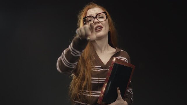 Redhead woman wearing glasses holding book makes l sign on forehead then points finger to camera in studio; mockery accusation.
