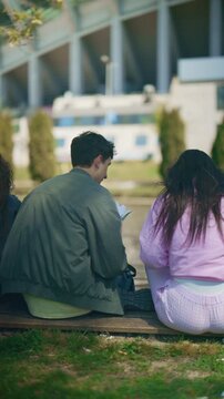 Two young high school students are sitting outdoors, discussing their classwork and sharing ideas. Classmates engage in a casual study session in a bright, sunny park environment.