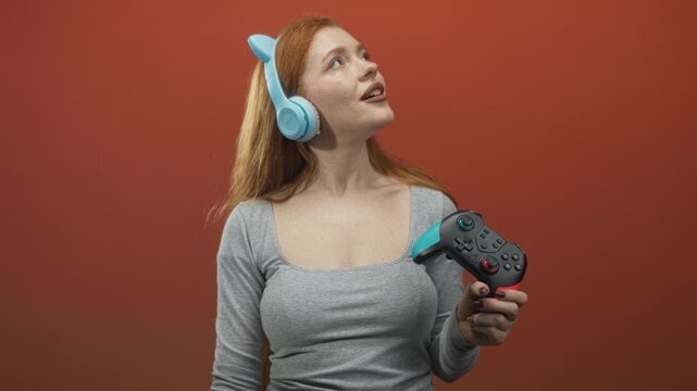 Woman holding a gamepad and wearing blue cat ear headphones, hand gripping a turquoise accented controller against a red studio wall; playful joy.
