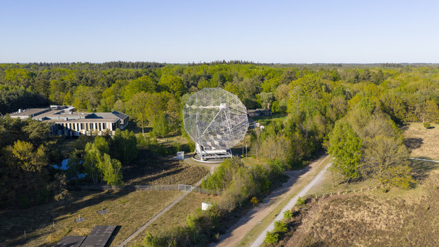 Aerial view of the Dwingeloo Radio Observatory dish surrounded by lush green forest and research buildings under a clear blue sky in Dwingeloo, Drenthe, Netherlands.