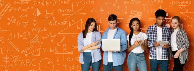 A group of young adults stands together in a learning area. They use laptops and phones to engage with their studies. Math and science notes are visible on the wall. © Prostock-studio