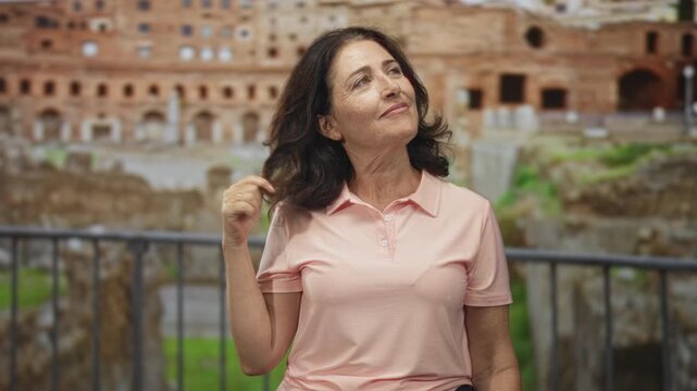 Woman gently twirls hair and smiles while looking up at ancient roman ruins and stone railing near a historic building; calm contentment.