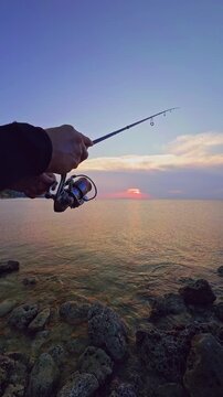 Vertical video. Hand Holding Fishing Rod Toward Sea Sunset First Person View Close Angle Outdoor Angling Action Horizon Reflection