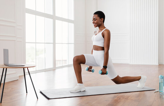 Sport And Healthy Lifestyle. Fit black woman watching training tutorial on laptop, doing forward walking lunges with dumbbells, exercising at home. Smiling lady looking at pc screen on tea table