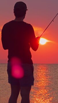 Vertical video. Man Silhouette Fishing Against Bright Sunset Over Sea Horizon Warm Light Reflection Coastal Outdoor Angling Lifestyle