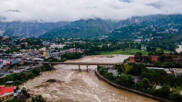 Aerial view of the urban landscape and a wide muddy river with a bridge nestled between lush green mountains and low clouds Muzaffarabad, Azad Kashmir, Pakistan.