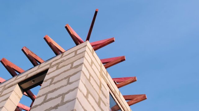 Low-angle view of unfinished house corner with white block walls and red treated wooden roof beams against clear blue sky, residential construction process and real estate development concept
