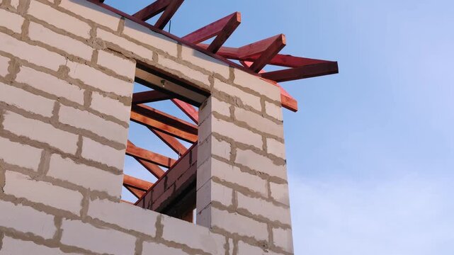 Low-angle view of unfinished house wall with window opening and red treated wooden roof frame, clear blue sky background, residential construction process and real estate development concept

