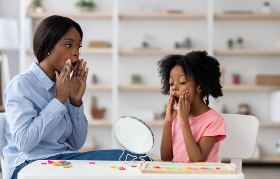 Young black woman speech-language pathologist exercising with adorable little girl with bushy hair, working on word sounds, kid looking at mirror and touching her mouth, clinic interior