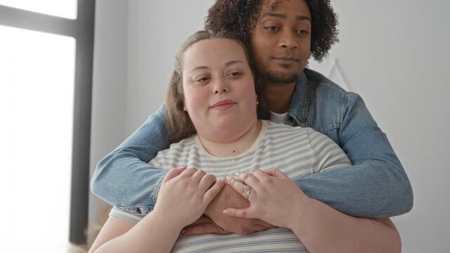 Woman embraced from behind by man, smiling, hands clasped over chest in studio; affection contentment.