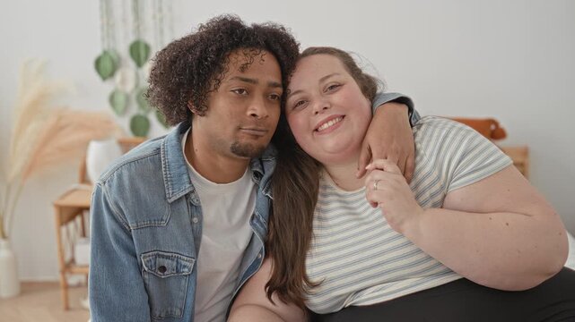Plus size woman sits on bed with arm around man, smiling toward camera, holding his hand and showing ring in a bright bedroom building; love together.