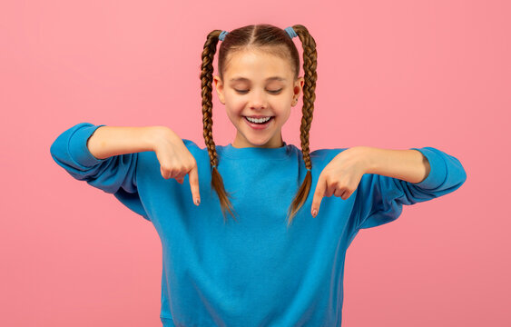 A cheerful young girl with two long braided pigtails is wearing a bright blue sweater. She stands against a solid pink backdrop, playfully pointing down with both her index fingers
