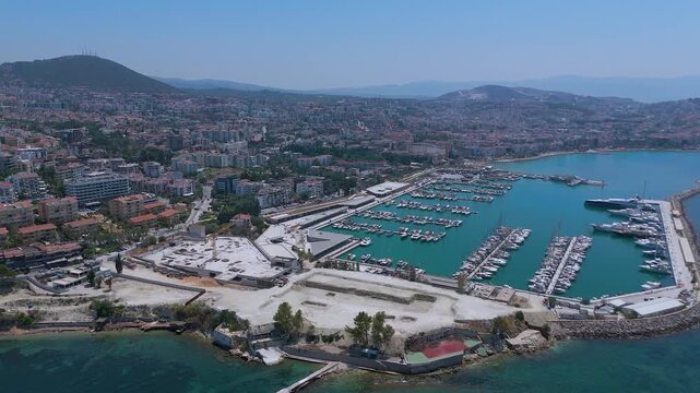 Kusadasi Marina in Turkey, showing rows of docked white yachts and sailboats in turquoise water, with a large cruise ship in the background and the coastal city sprawling across the hills.