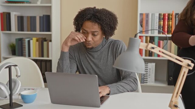Man at laptop touching ear with finger beside tall stack of files on desk in studio; quiet concern focus.