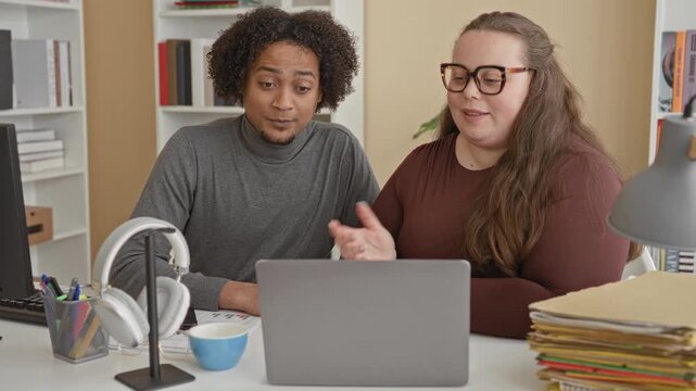 Man and woman sit at desk, man pointing hand to laptop among stacks of folders and headphones in an office building; teamwork focused.