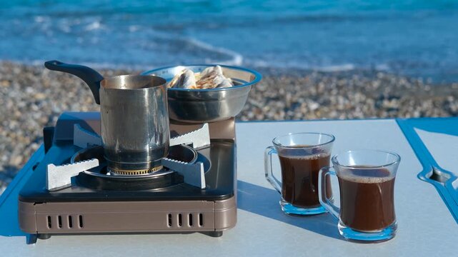 Camping coffee making on beach with portable stove. Portable gas stove preparing coffee in cezve near two cups of coffee bowl on folding table on pebble beach near sea waves on sunny summer day