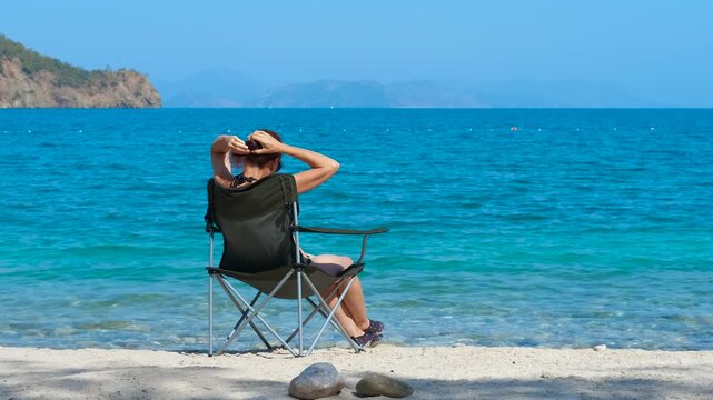Woman sitting on beach chair, gently fixing windblown hair while experiencing peaceful seaside moment, embodying serene leisure and personal tranquility