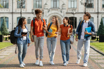 Fototapeta premium Happy university students walking together on campus, group of cheerful young multiethnic people chatting and laughing outdoors during break, carrying backpacks and workbooks, full length shot