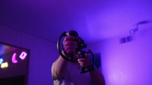 Man in VR headset pointing controllers forward actively playing at home performing movements and interacting with virtual environment immersing into gameplay under neon lighting