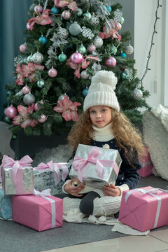 Cute little girl in winter clothes posing at home front of Christmas tree with gift box in hand. Christmas and New year concept. Holiday home decoration. Selective focus