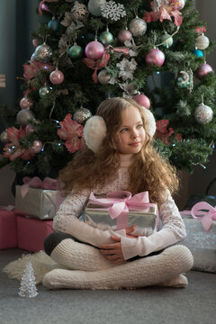 Cute little girl in winter clothes posing at home front of Christmas tree with gift box in hand. Christmas and New year concept. Holiday home decoration. Selective focus