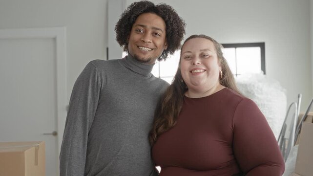 Man in grey turtleneck with arm around curvy woman in maroon top waving hand beside stacked moving boxes in a building interior; love together joy home.