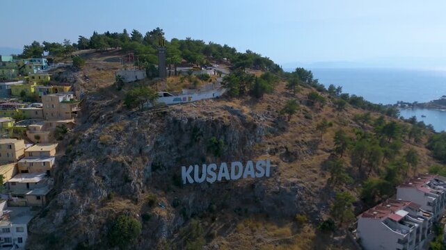 Aerial view of the iconic white "KUSADASI" sign on a rocky cliffside, featuring the Ataturk Hill (Ataturk Tepesi) with the golden statue of Mustafa Kemal Ataturk at the summit.