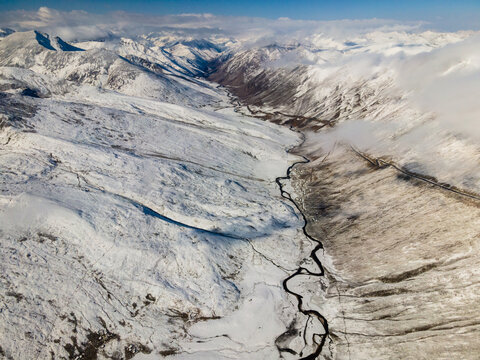 Aerial view of Babusar Pass with snow-covered mountain peaks, a winding road through a deep valley, and thick white clouds under a clear blue sky Babusar, KPK, Pakistan.