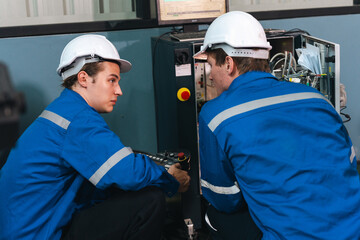 Two male engineers wearing blue uniforms and white safety helmets are working together on industrial equipment, focusing on maintenance and repair in modern factory setting