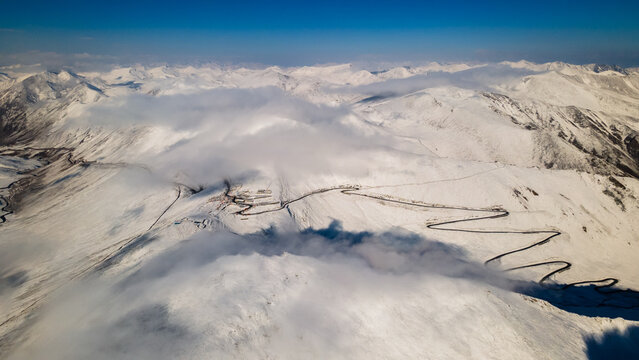 Aerial view of Babusar Pass with its winding road snaking through snow-covered mountain peaks under a clear blue sky and soft clouds in Babusar, KPK, Pakistan.