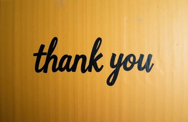 Naklejka premium Black script text saying thank you printed on rough yellow cardboard. Simple appreciation message on textured box surface, copy space available for design.