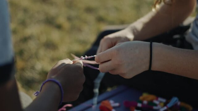 Two friends weaving colorful threads to create handmade friendship bracelets together outdoors