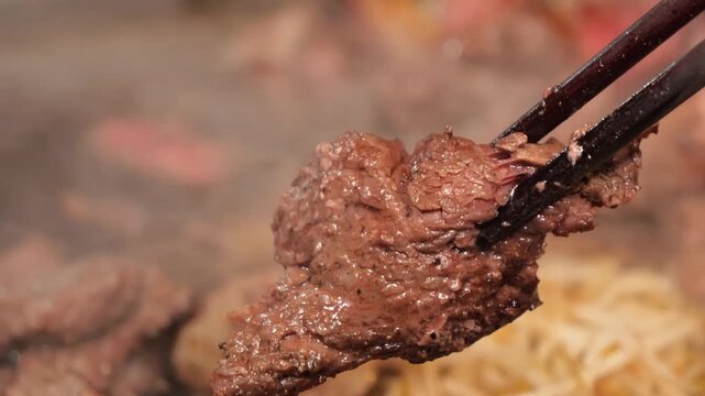 A high-quality close-up shot of tender, well-marinated beef pieces being lifted with black chopsticks. The meat is perfectly grilled, showing a succulent, juicy texture with visible steam, capturing t