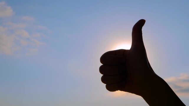 Close up shot: silhouette of woman is showing thumbs up hand gesture against the sunset, sunrise sky with sun lens flares. Approval, satisfaction and positive concept