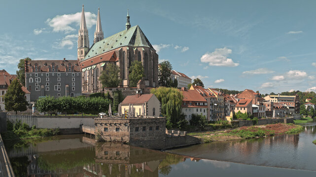 Historic European cityscape featuring a grand Gothic church with twin spires towering over charming old houses, a calm river with reflections, and a stone weir under a sunny sky.