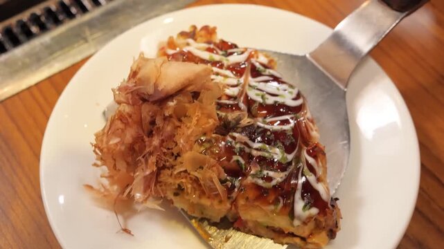 close-up shot of a freshly prepared Osaka-style okonomiyaki (Japanese savory pancake) topped with thick brown sauce, mayonnaise, and dancing bonito flakes (katsuobushi). The footage shows a slice bein