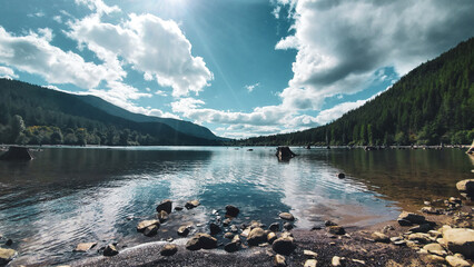 Sunbeams Piercing Through Clouds Over a Serene Mountain Lake with Stumps and Rocky Shoreline © Nick