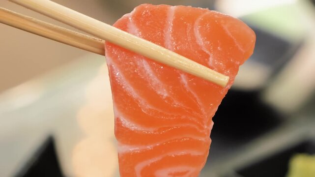 A slow-motion closeup of a delicate, buttery slice of fresh salmon sashimi being picked up with wooden chopsticks. The footage highlights the vibrant orange color and white marbling of the premium, ra