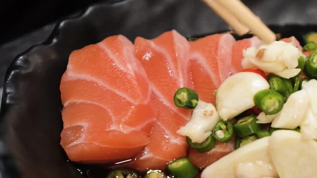 A high-angle, close-up shot of vibrant, thinly sliced raw salmon sashimi marinated in a savory and spicy Thai sauce. The dish is beautifully garnished with fresh green and red chilies and thin slices 