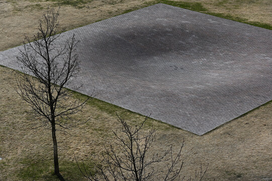A minimalist aerial-style photograph of a geometric brick-paved square set within a sparse park landscape