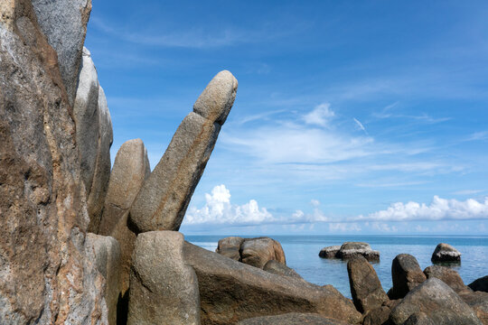 Granite rock formation resembling a phallic shape rising against blue sky with clouds on tropical island coastal landscape.
