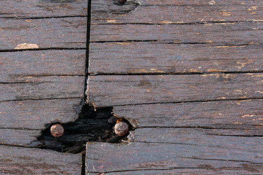 Aged wooden planks with visible grain, cracks, and a damaged section exposing inner wood, featuring two rusty nails embedded in the surface.

